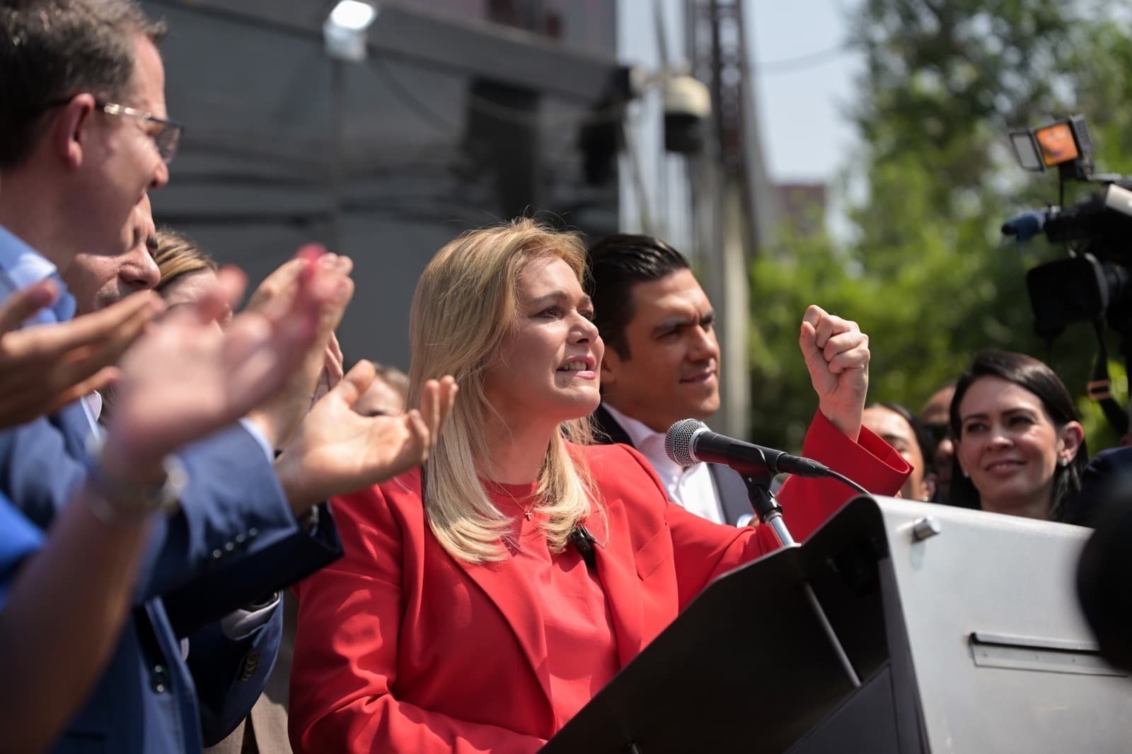 La candidata política pronuncia un discurso en un evento, rodeada de simpatizantes que la aplauden y muestran apoyo.