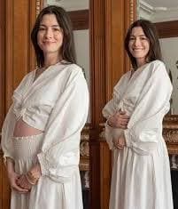 Anne Hathaway sonríe mientras muestra su avanzado embarazo, vestida con una blusa blanca, en un ambiente acogedor.