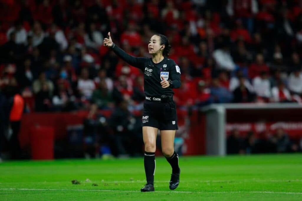 Katia Itzel García, árbitra mexicana, dirigiendo un partido con gesto de aprobación en un estadio lleno de aficionados.