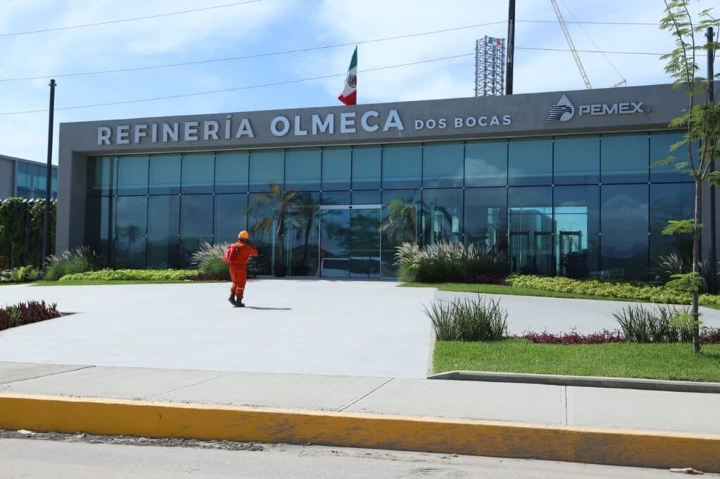 Trabajador caminando hacia la entrada de la Refinería Olmeca en Dos Bocas, Tabasco, con bandera mexicana ondeando.