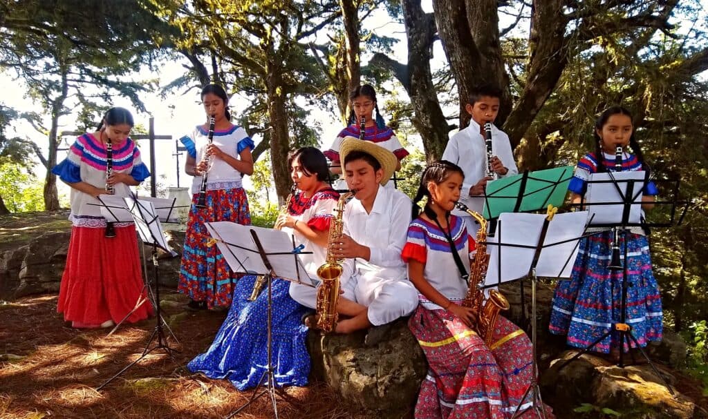 Niños y jóvenes vestidos con trajes tradicionales tocan instrumentos musicales en un entorno natural, promoviendo la