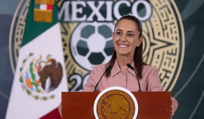 Claudia Sheinbaum Pardo sonríe mientras entrega boletos del Mundial 2026 en conferencia, con bandera mexicana de fondo.