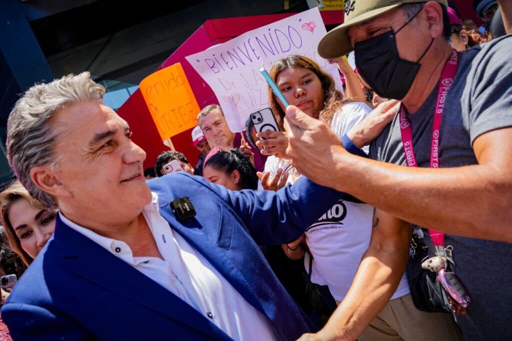 Jesús Alejandro Ruiz Uribe saluda a simpatizantes en un evento de MORENA en Baja California, mientras sostienen pancartas de apoyo.