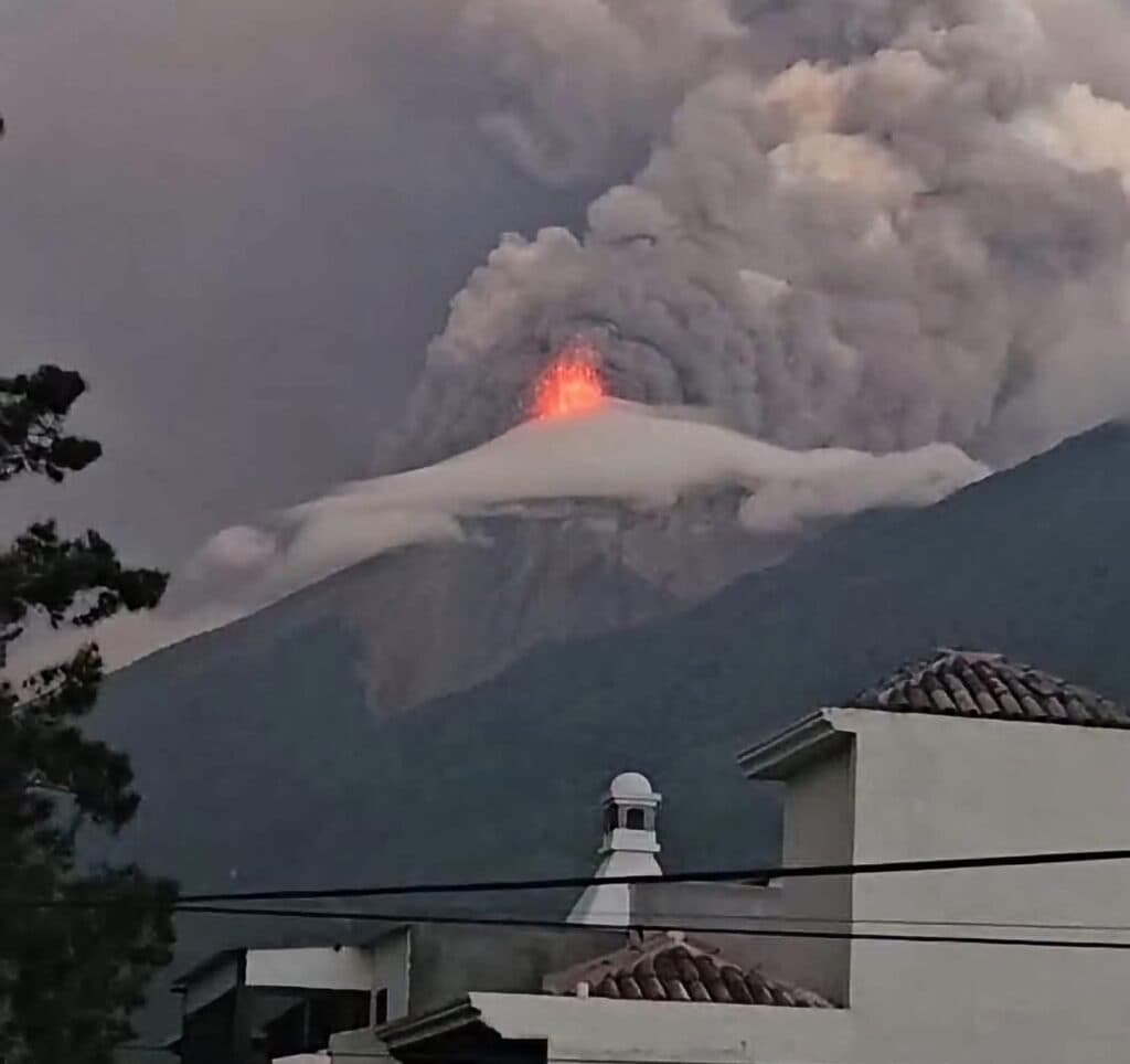 Erupción del Volcán de Fuego en Guatemala, con emisión de lava y nubes de ceniza, visible desde una zona residencial cercana.