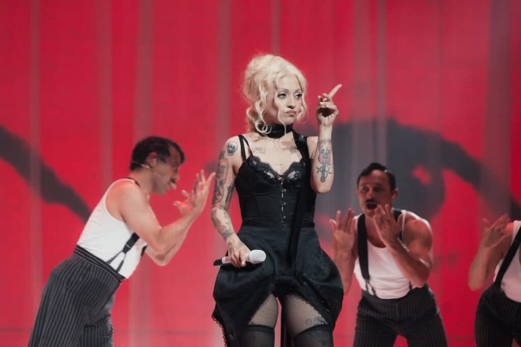 Mon Laferte en el escenario durante una presentación, luciendo un vestido negro y rodeada de bailarines con trajes a rayas.