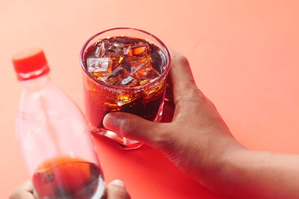 Vaso con refresco oscuro y hielo, mientras una mano sostiene una botella de bebida azucarada, destacando el riesgo para la salud hepática.