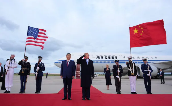 Xi Jinping y Donald Trump en la base aérea Andrews, saludando con banderas de Estados Unidos y China en el contexto de una recepción oficial.