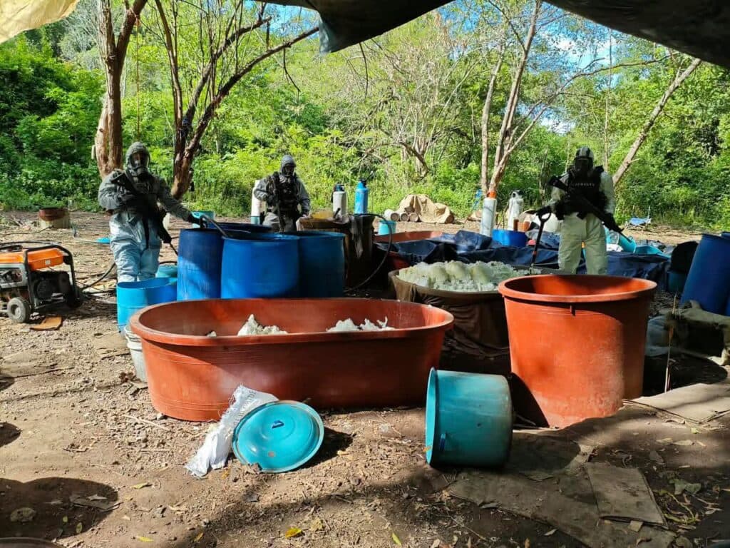 Autoridades federales y estatales en un laboratorio clandestino desmantelado en Michoacán, rodeados de barriles y equipos de