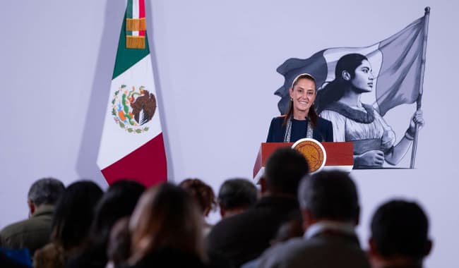 Claudia Sheinbaum en conferencia de prensa en Palacio Nacional, con bandera de México y mural de mujer indígena al fondo.
