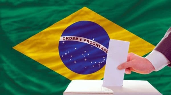 Una mano deposita un voto en una urna frente a la bandera de Brasil, simbolizando la elección presidencial entre Lula y Flávio Bolsonaro.