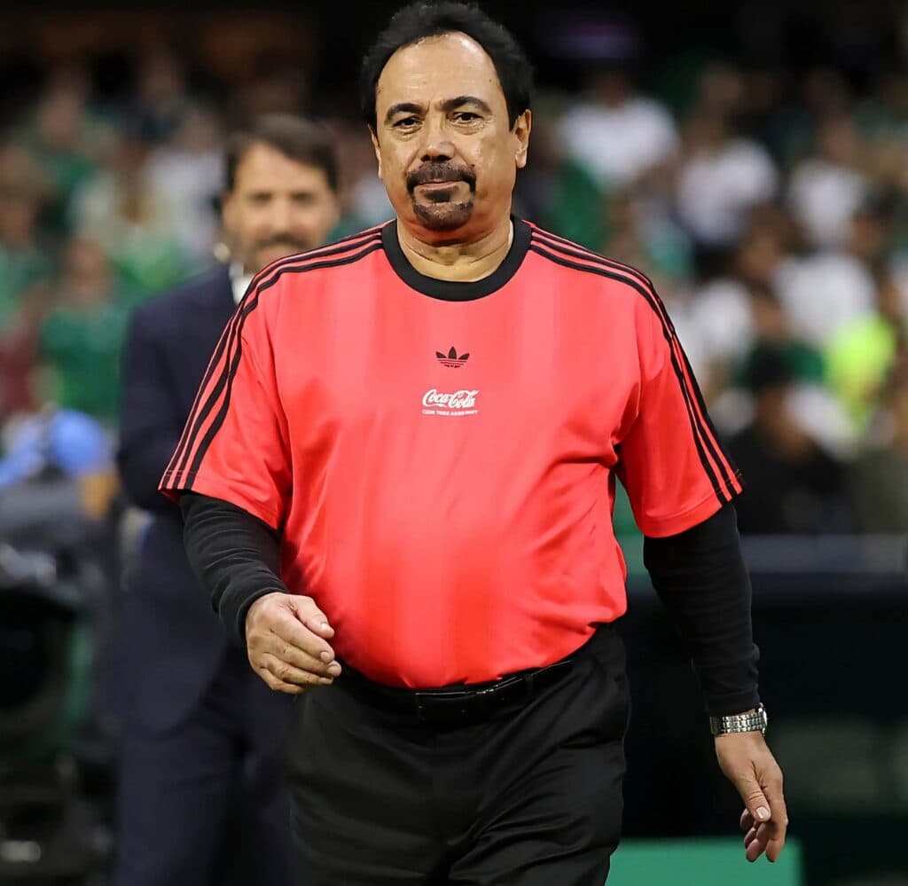 Hugo Sánchez, exfutbolista mexicano, vestido con camiseta roja de árbitro, camina por el campo durante un evento deportivo.