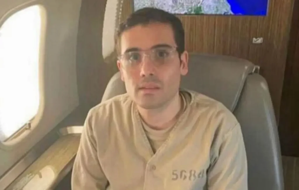 Ovidio Guzmán, hijo de Joaquín "El Chapo" Guzmán, sentado en un avión privado, vestido con uniforme carcelario, en contexto